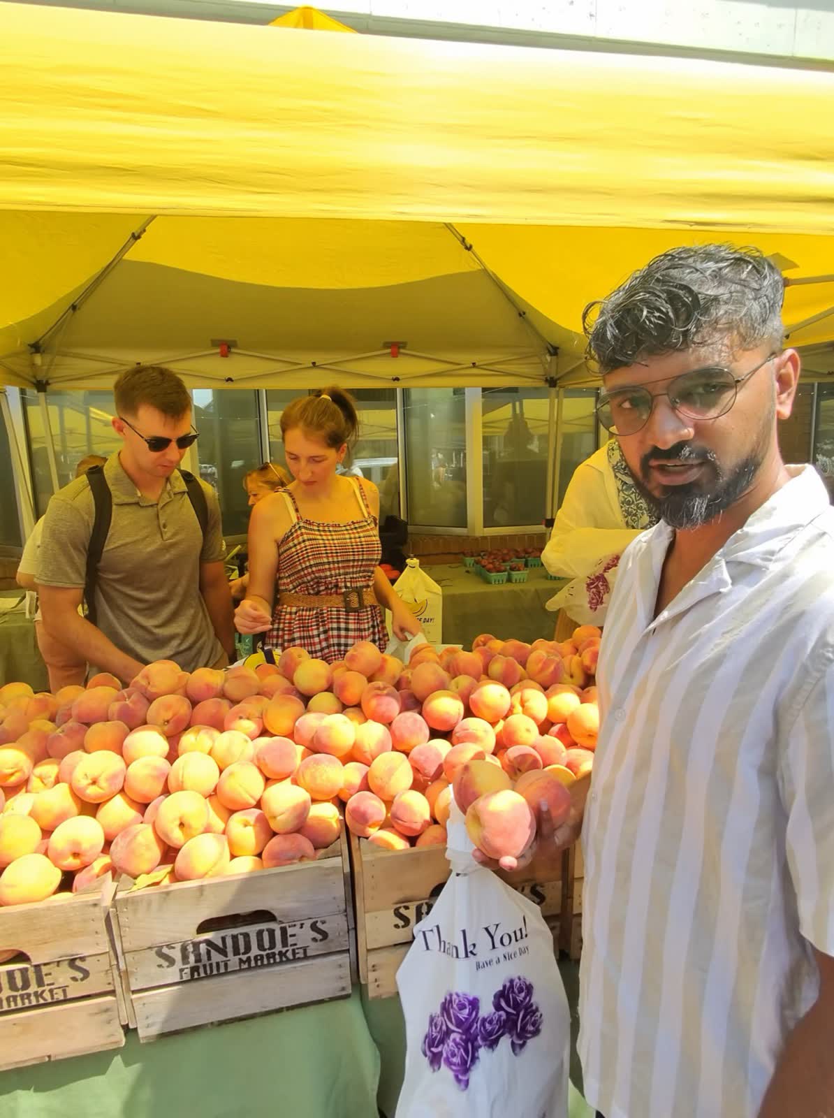 A vendor under a yellow tent stands behind crates of fresh peaches at a Saturday farmers market